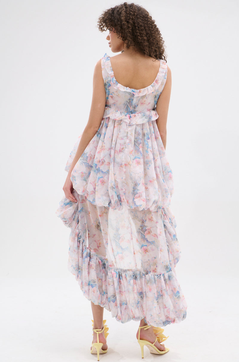 A woman with curly hair stands with her back to the camera in the sleeveless, sheer floral THROUGH THE MEADOW RUFFLE HIGH LOW DRESS and yellow high-heeled sandals against a white background.