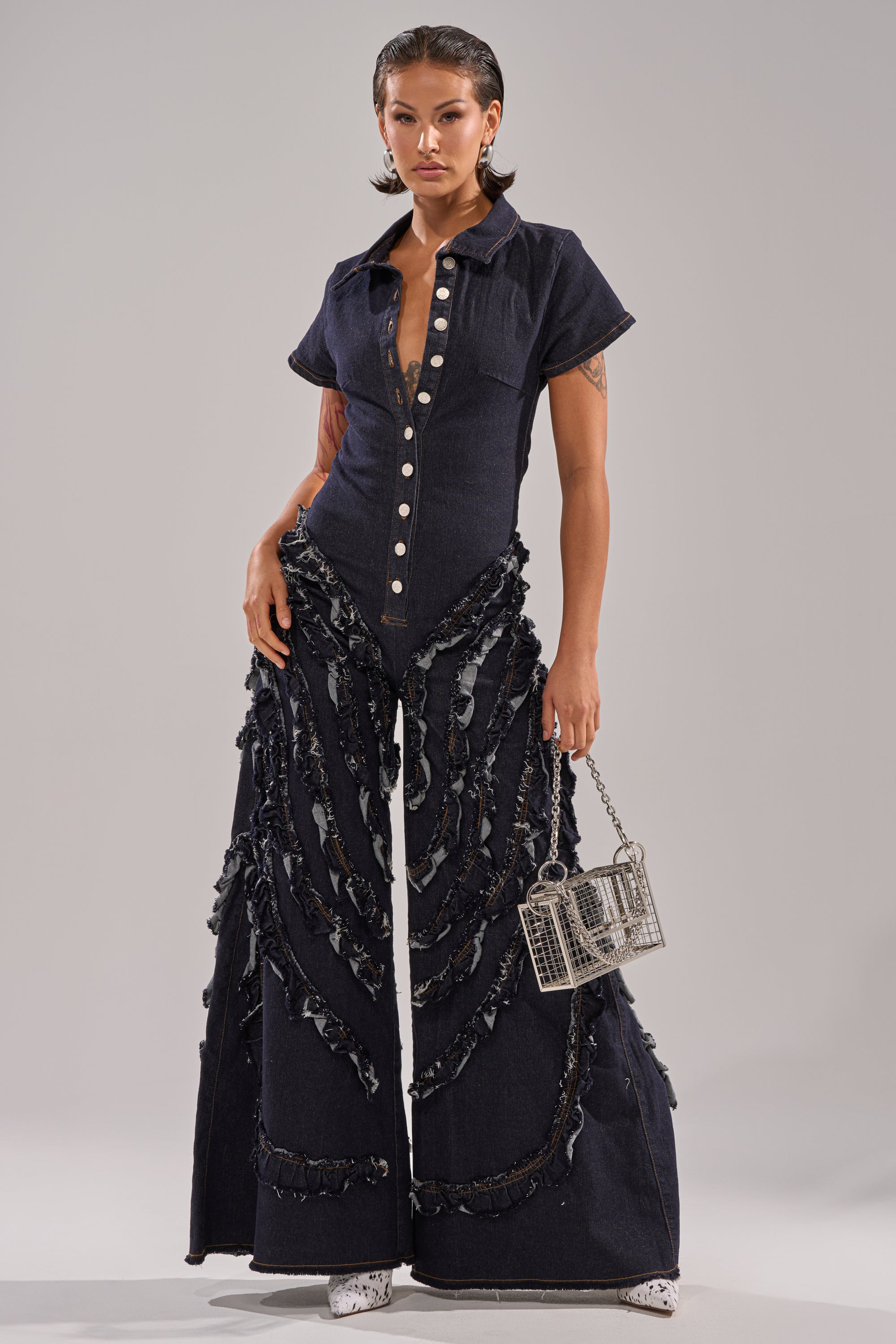 A person stands confidently in the INTO THE NIGHT DENIM JUMPSUIT, featuring dark wash denim with wide flared legs and frayed details, styled with a silver cage handbag and white boots against a light gray background.