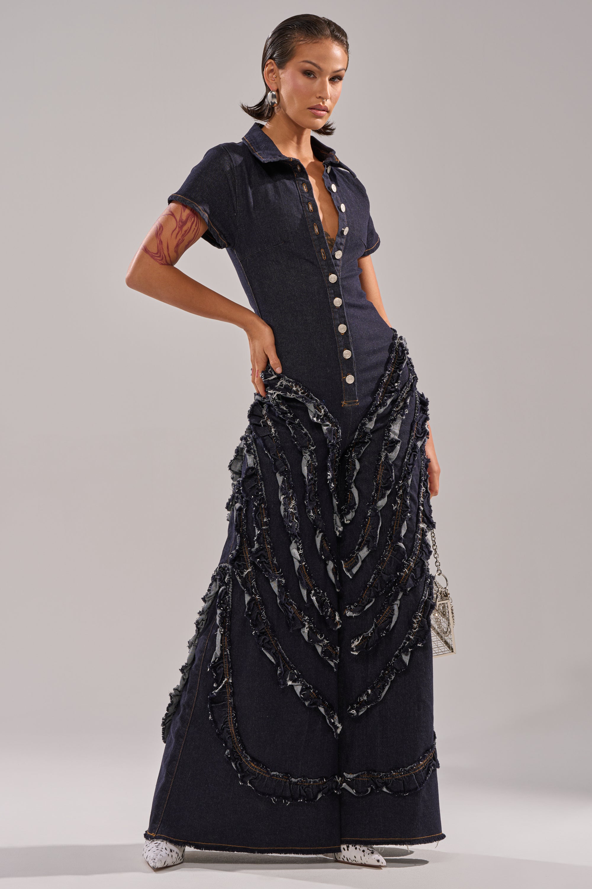 A model wears the INTO THE NIGHT DENIM JUMPSUIT, featuring a dark wash, short sleeves, collared neckline, bold silver front buttons, and wide-leg pants with layered textured details. Her hair is slicked back for a sleek look.