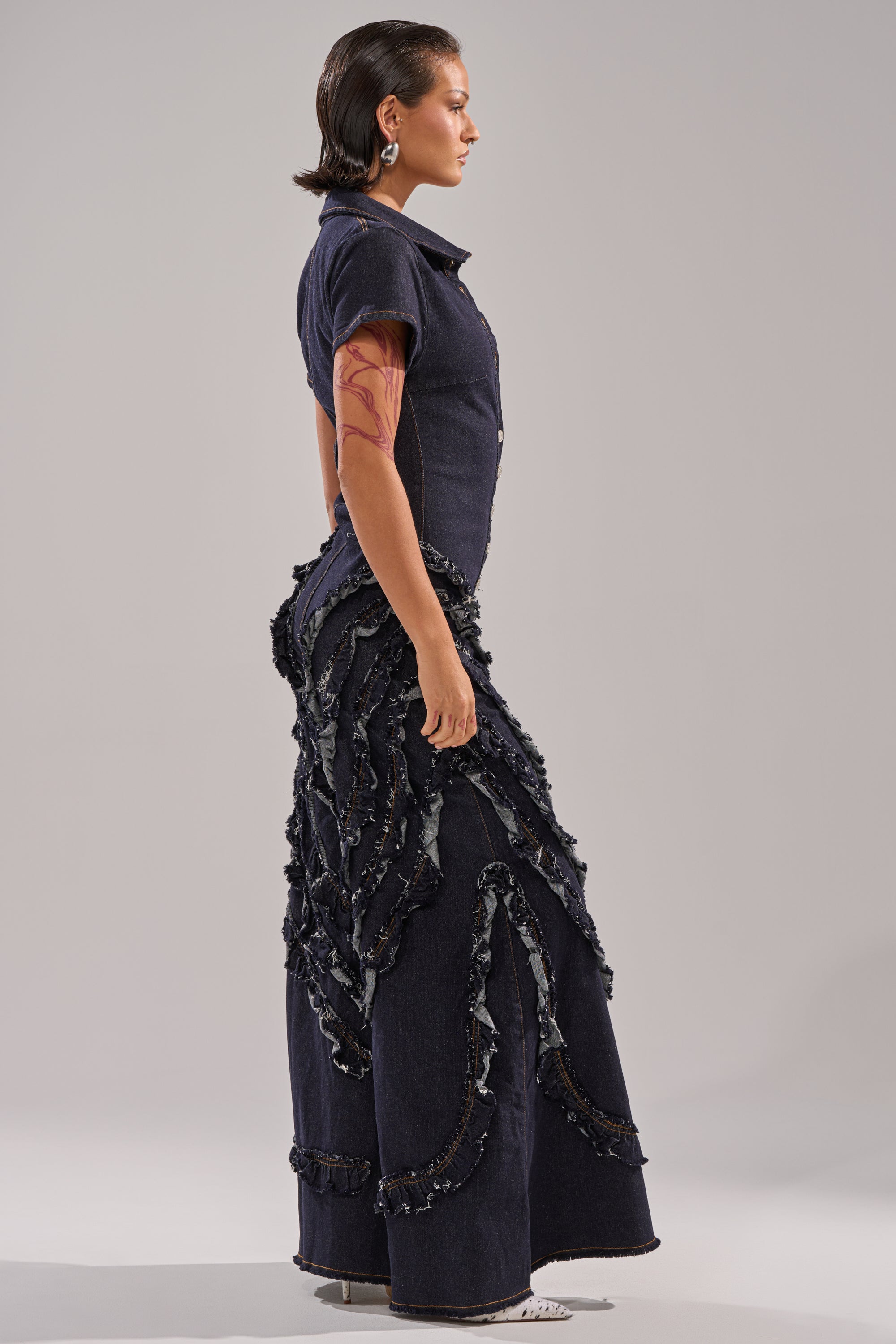 A person in profile wears the INTO THE NIGHT DENIM JUMPSUIT, featuring a dark wash, short sleeves, wide-leg pants, and wavy frayed accents. Their hair is slicked back and an arm tattoo is visible.