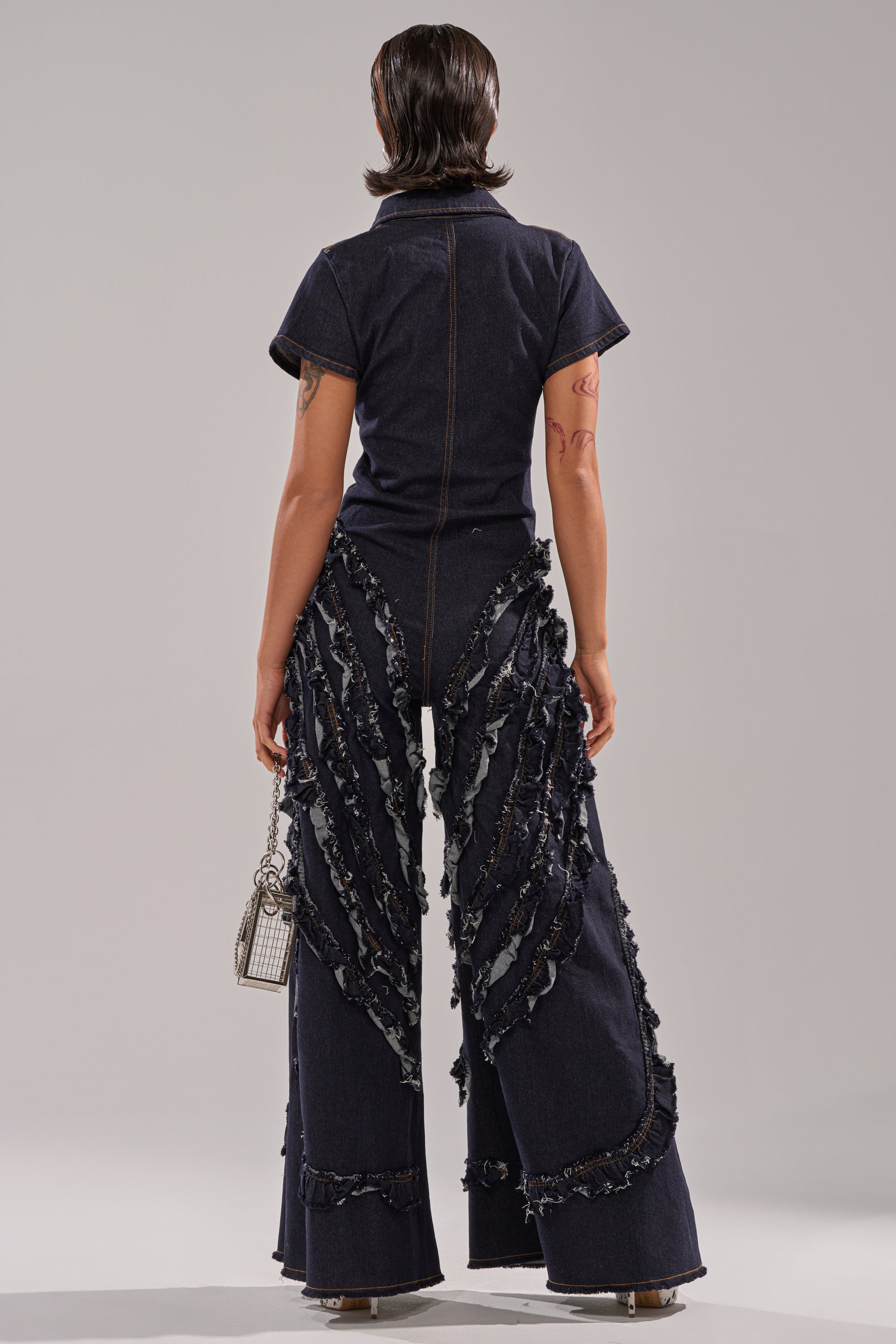 A person faces away, wearing the INTO THE NIGHT DENIM JUMPSUIT—dark-wash with short sleeves, wide-leg pants, and layered frayed strips. They carry a metallic handbag and have short, styled hair.