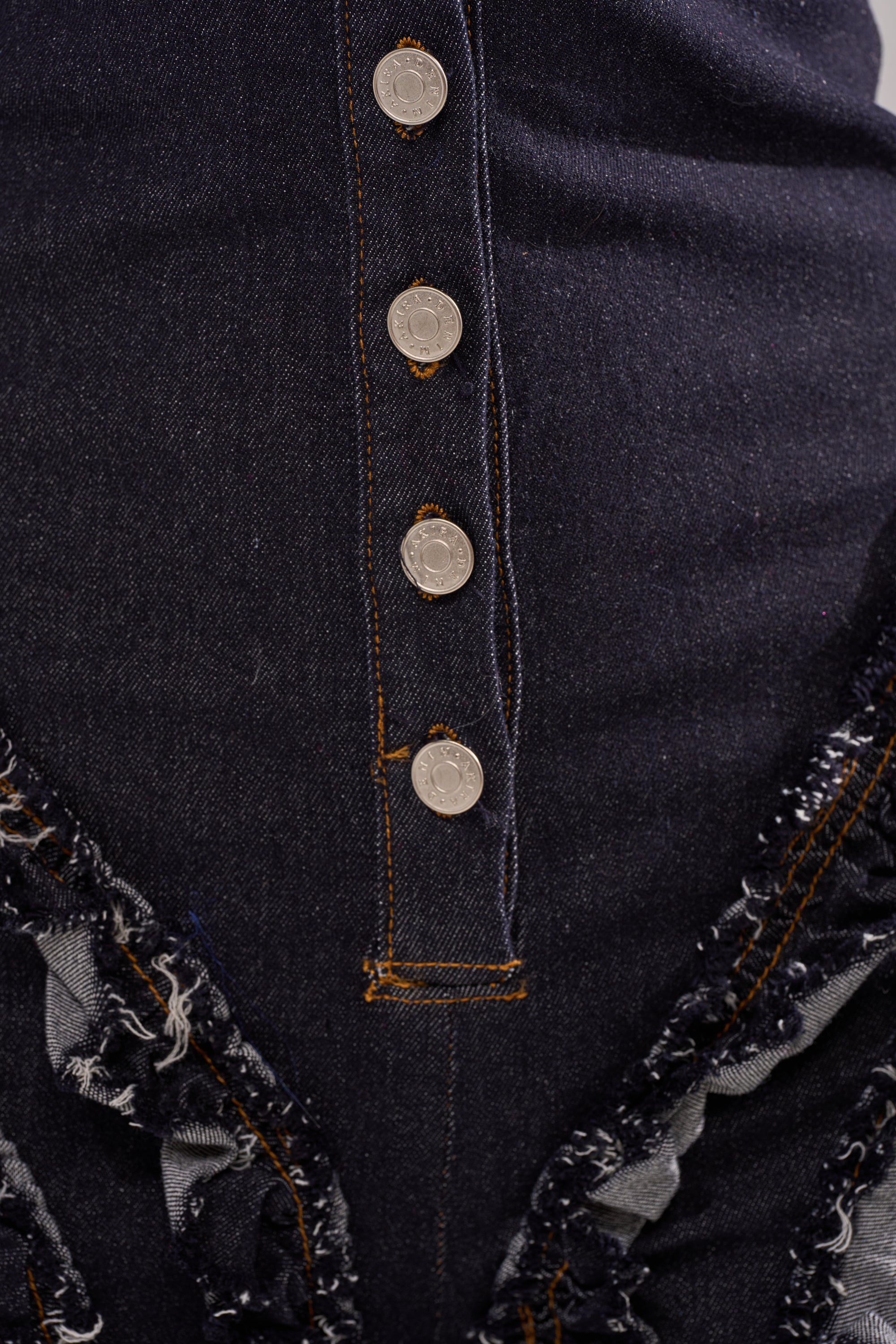 Close-up of the INTO THE NIGHT DENIM JUMPSUIT in dark blue denim with a vertical row of four silver buttons, visible orange stitching, distressed ruffle accents, and layered denim details on each side.