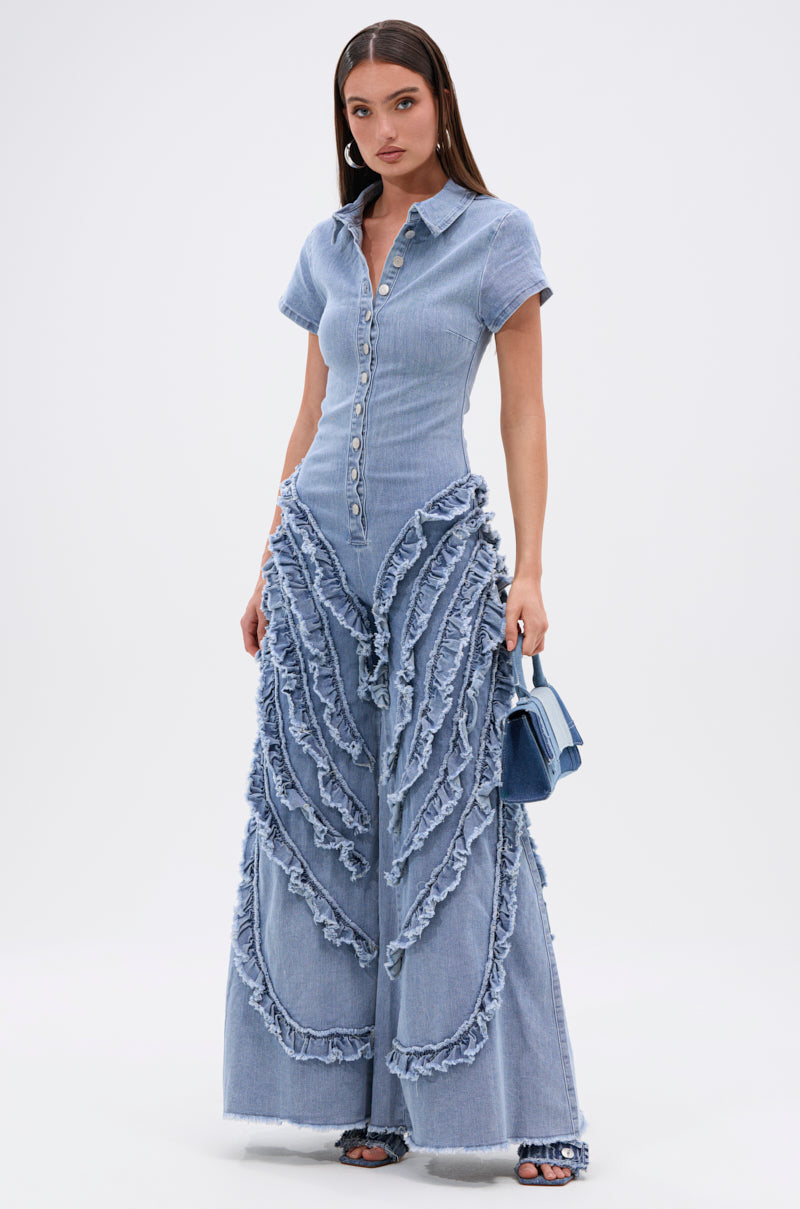 A woman models the LEAVE IT RUFFLED JUMPSUIT IN DENIM, featuring light blue denim, short sleeves, button-down front, wide-leg pants with bold wavy frayed accents. She carries a blue handbag and wears open-toe shoes to highlight this trendy look.