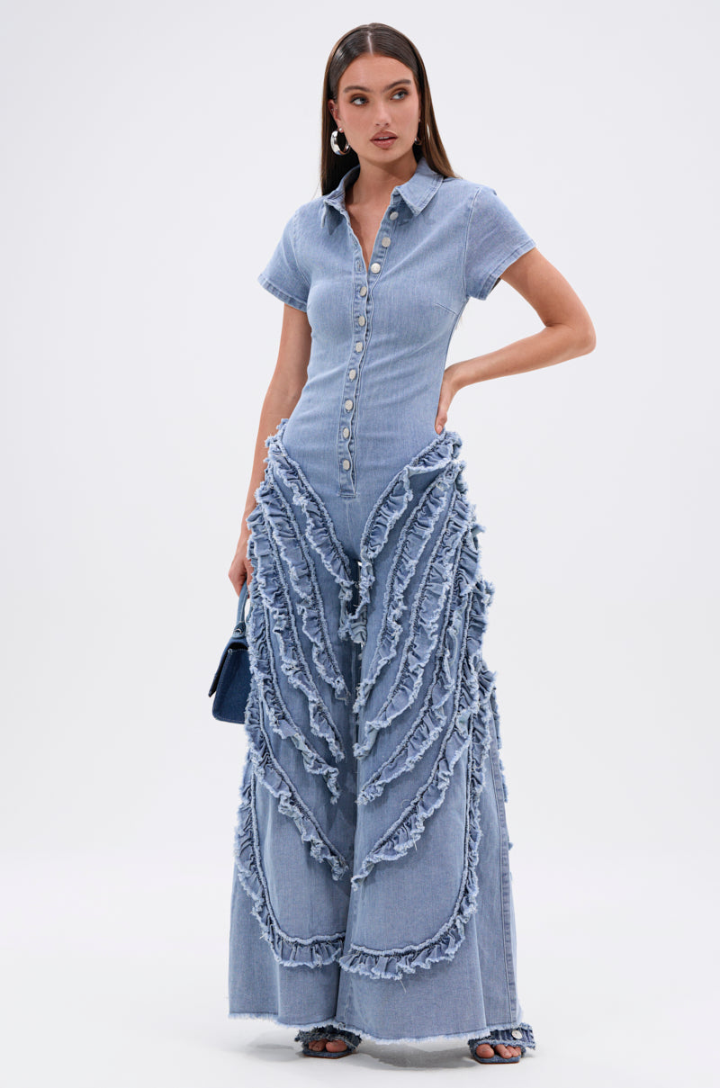 A woman models the LEAVE IT RUFFLED JUMPSUIT IN DENIM, featuring short sleeves, a buttoned front, and wide-leg layered ruffles. She accessorizes with a navy bag, open-toe heels, and statement earrings.