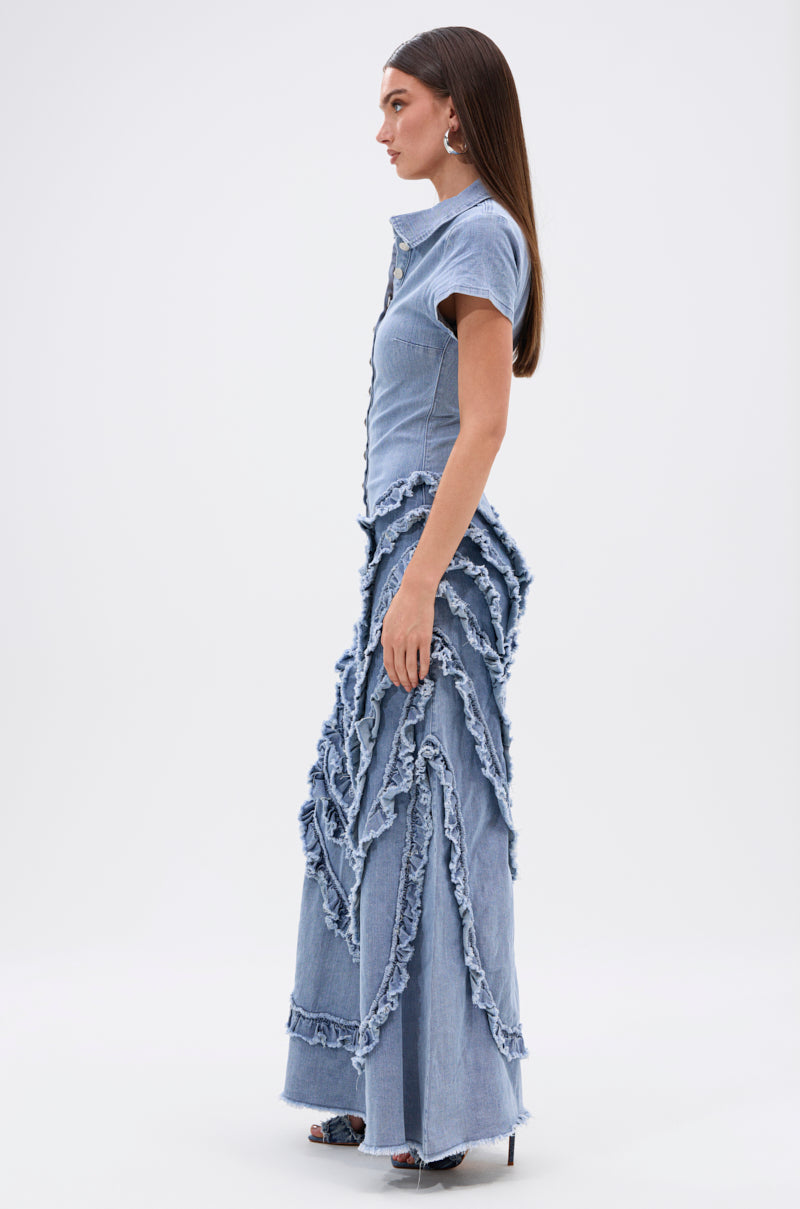 Woman standing in profile against a plain background, wearing the LEAVE IT RUFFLED JUMPSUIT IN DENIM—a short-sleeved, form-fitting denim jumpsuit with wavy, frayed trim evoking wide-leg pants. She has straight, long hair and heeled sandals.