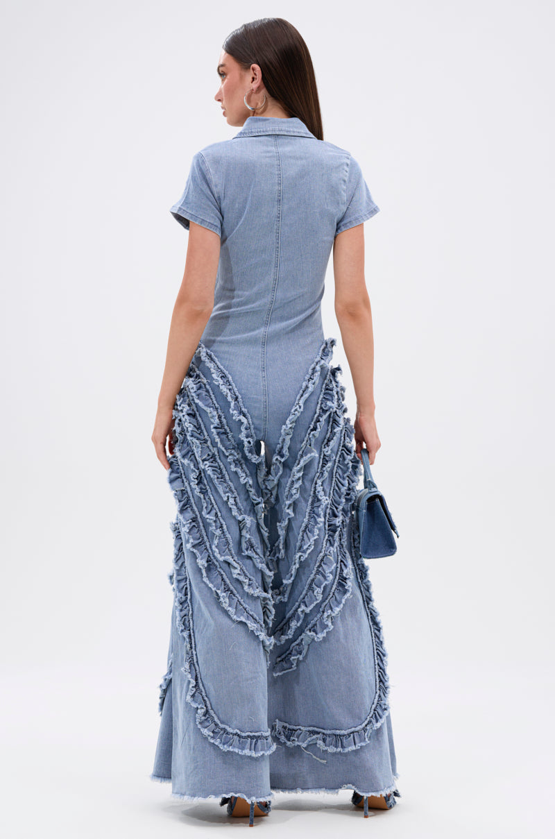 A woman models the LEAVE IT RUFFLED JUMPSUIT IN DENIM, featuring light blue denim with short sleeves, wide-leg pants, and cascading ruffles on the back. She accessorizes with a small denim handbag and heeled sandals.