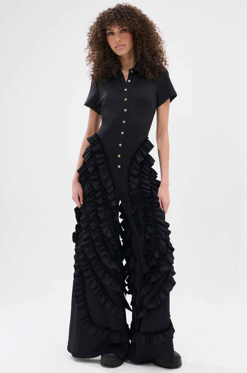 A woman with curly hair models the LEAVE IT RUFFLED JUMPSUIT IN BLACK, showcasing gold buttons, dramatic ruffled details along the sides and legs, and a wide-leg silhouette against a plain white background.