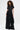 A woman with curly hair models the LEAVE IT RUFFLED JUMPSUIT IN BLACK, showcasing gold buttons, dramatic ruffled details along the sides and legs, and a wide-leg silhouette against a plain white background.