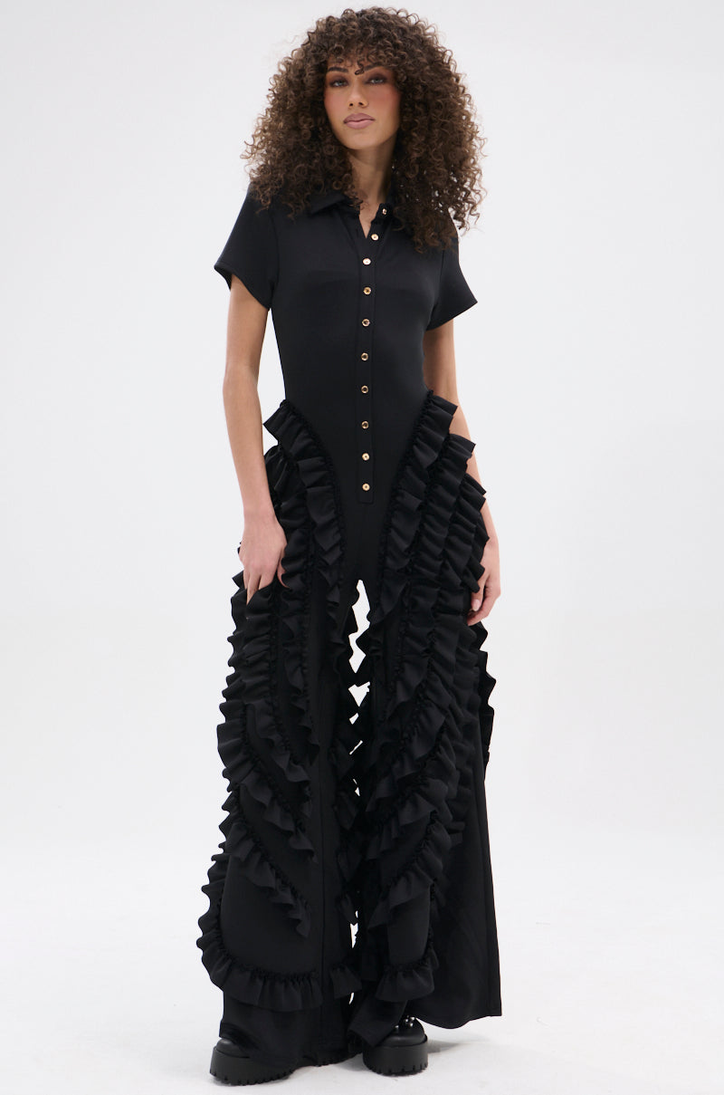 A woman with curly hair wears the LEAVE IT RUFFLED JUMPSUIT IN BLACK, made from stretchy fabric with short sleeves and bold vertical ruffles down wide legs, standing against a plain white background.