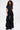 A woman with curly hair wears the LEAVE IT RUFFLED JUMPSUIT IN BLACK, made from stretchy fabric with short sleeves and bold vertical ruffles down wide legs, standing against a plain white background.