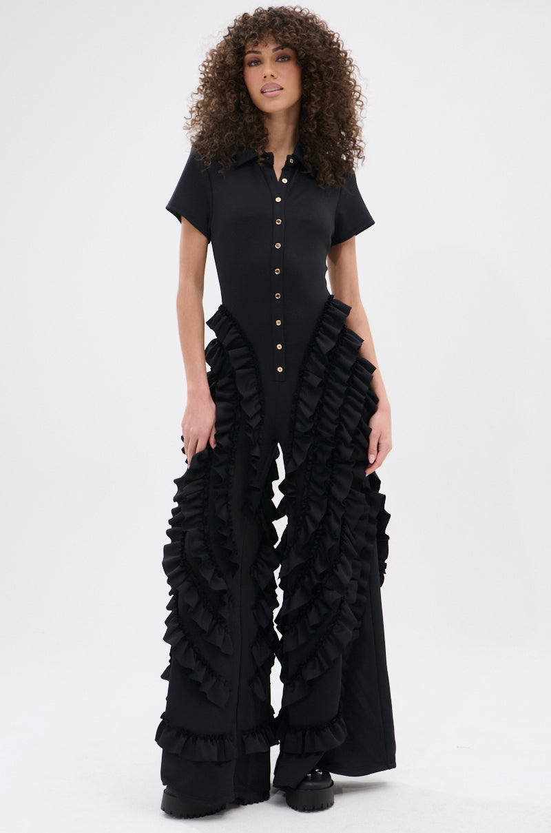 A woman with curly hair models the LEAVE IT RUFFLED JUMPSUIT IN BLACK, featuring short sleeves, a button-up front, and dramatic wide-leg style, standing against a plain white background.