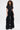 A woman with curly hair models the LEAVE IT RUFFLED JUMPSUIT IN BLACK, featuring short sleeves, a button-up front, and dramatic wide-leg style, standing against a plain white background.