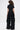 A person with curly hair models the LEAVE IT RUFFLED JUMPSUIT IN BLACK, showcasing its short sleeves and dramatic ruffled details cascading down the wide-leg silhouette. The photo is shot from behind against a plain white background.