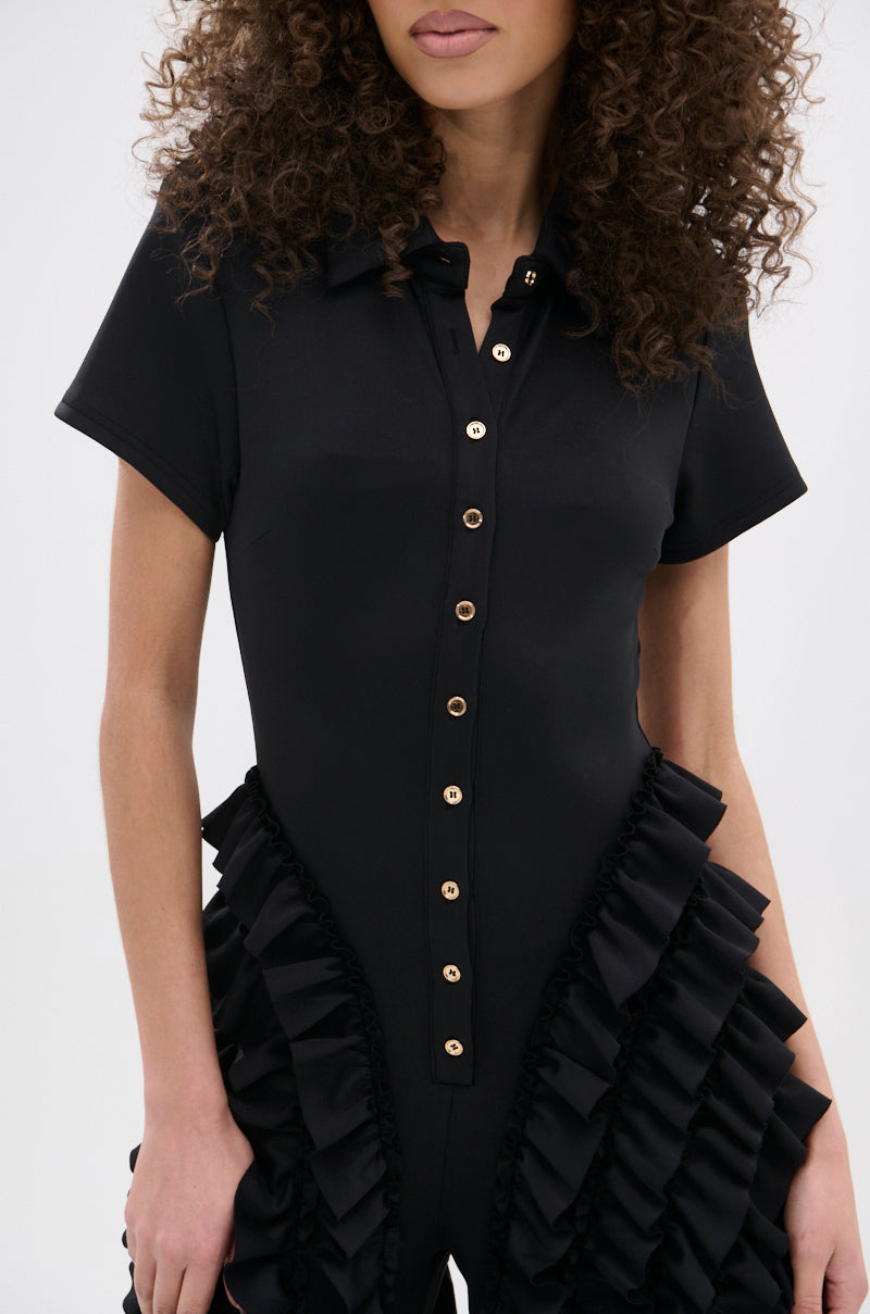 A woman with curly hair models the LEAVE IT RUFFLED JUMPSUIT IN BLACK, featuring gold buttons and ruffled side details. The cropped photo highlights its chic wide-leg silhouette from chest to mid-thigh.