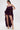A woman confidently poses in ROYAL BLOOMER SHORTS, styled under a ruffled sheer deep purple high-low dress with spaghetti straps. She completes the look with gold platform heels, large hoop earrings, and a sleek ponytail.
