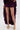 A person stands against a plain white background, wearing ROYAL BLOOMER SHORTS under a sheer, ruffled deep purple high-low skirt and gold platform heels, gently holding the sides of the skirt.