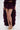 A person wears the ROYAL BLOOMER SHORTS, featuring a sheer, ruffled, dark purple design with a high slit and high-rise fit, styled with gold platform sandals. The background is plain white.