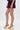 A person wearing ROYAL BLOOMER SHORTS in dark purple and shiny gold platform heels stands against a plain white background.