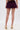 A person stands on a white surface, shown from the waist down, wearing ROYAL BLOOMER SHORTS in a high-rise sheer chiffon style paired with gold high-heeled sandals featuring ankle straps.