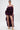 A woman stands facing forward in a sheer, deep purple ruffled two-piece featuring a crop top and ROYAL BLOOMER SHORTS. The high-rise fit and long train are paired with hoop earrings, gold platform heels, and her hair pulled back.