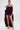 A woman stands confidently against a plain white background, wearing chic ROYAL BLOOMER SHORTS styled with gold platform heels and a layered, ruffled, dark purple top featuring spaghetti straps for a trendy high-rise look.