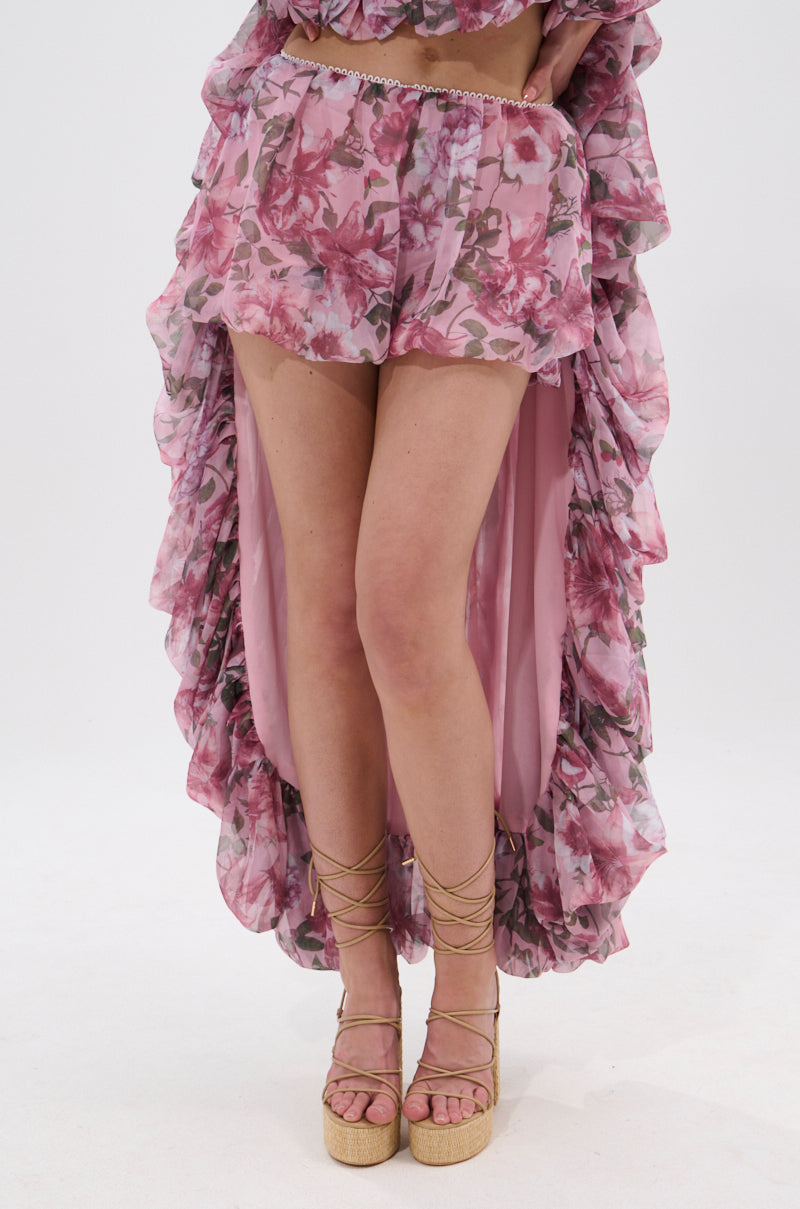 A person wears REVERENT FLORAL BLOOMER SHORTS with a pink floral high-low ruffled skirt and beige lace-up platform sandals, all in lightweight chiffon, set against a plain white background.