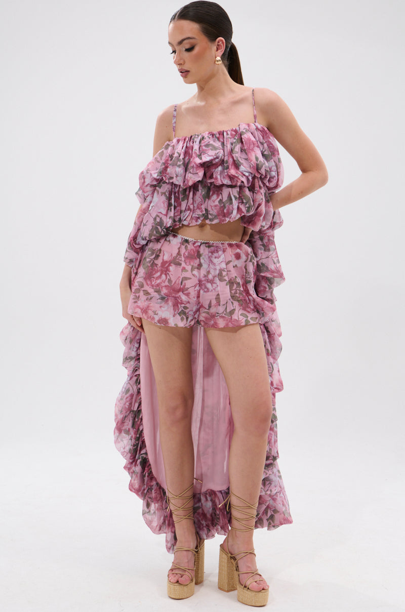 Woman in REVERENT FLORAL BLOOMER SHORTS—a pink, high-waisted set with ruffled off-shoulder crop top and flowing train. Her hair is tied back and she finishes the look with strappy wedge sandals.