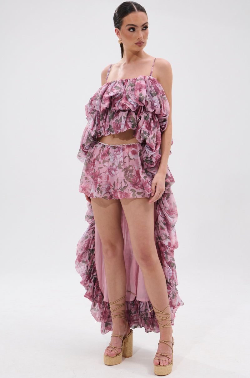 A woman wears AKIRA Label's REVERENT FLORAL BLOOMER SHORTS—a pink floral, high-waisted, ruffled two-piece set with a spaghetti strap crop top and lace-up wedge sandals.