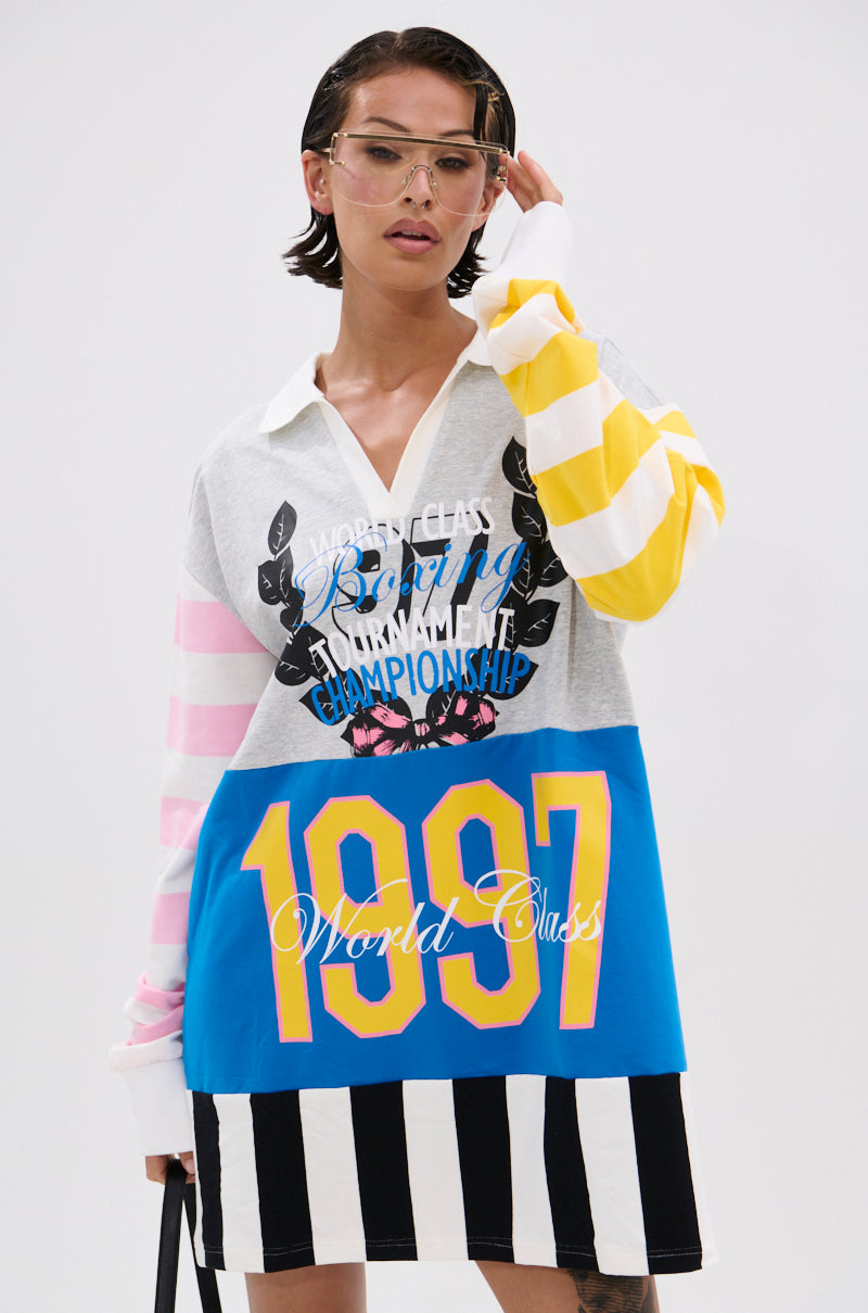 A person with short hair wears the HERE FOR THE DRINKS PRINTED TSHIRT DRESS—a colorful, oversized T-shirt dress with varsity graphics, striped sleeves in yellow and pink, colorblock stripes at the bottom, and large glasses.