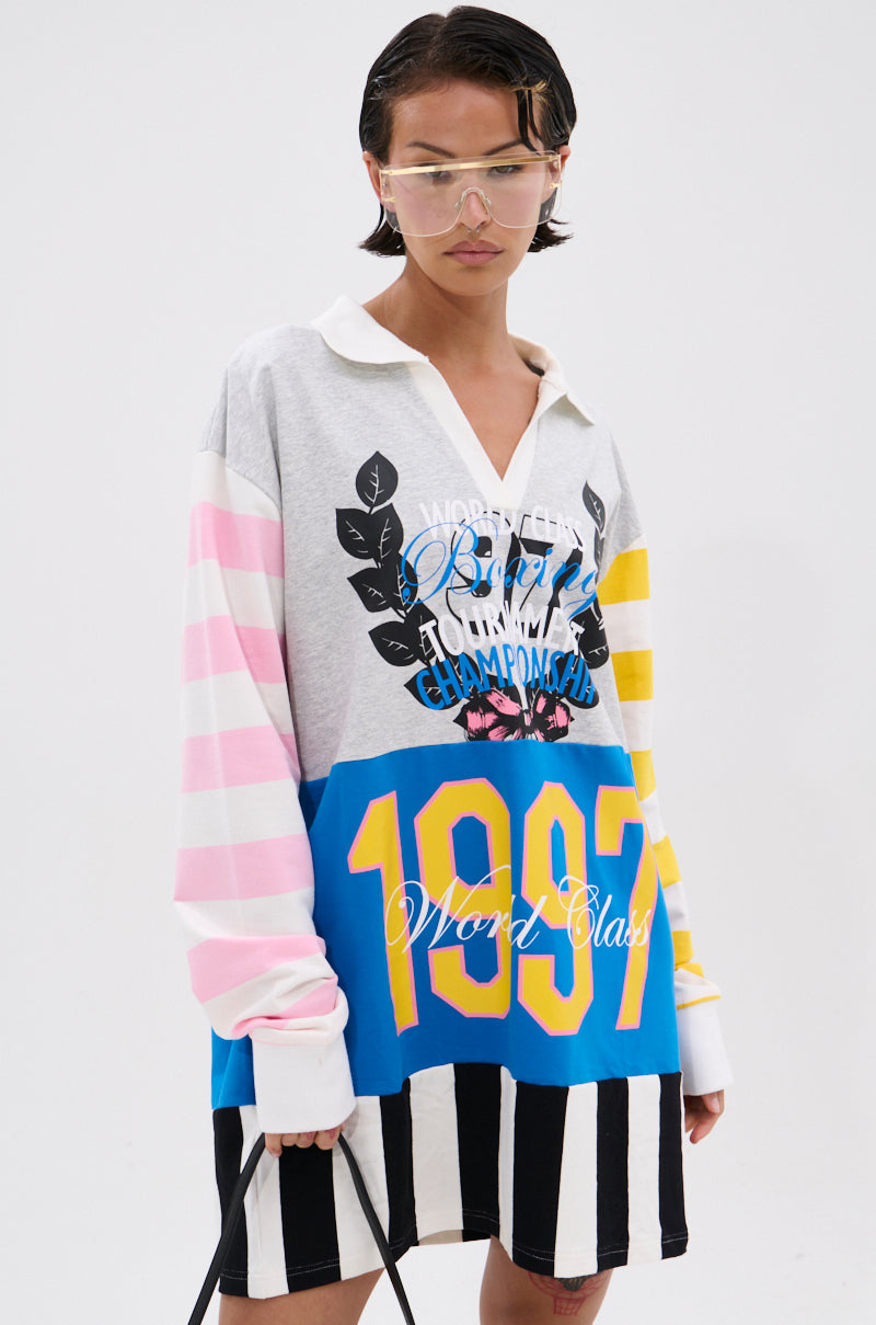 A person with short hair and clear visor glasses wears the HERE FOR THE DRINKS PRINTED TSHIRT DRESS—an oversized, colorful dress with mixed patterns, colorblock stripes in pink, yellow, blue, black-and-white, and varsity-style "1997" and "World Class" graphics.
