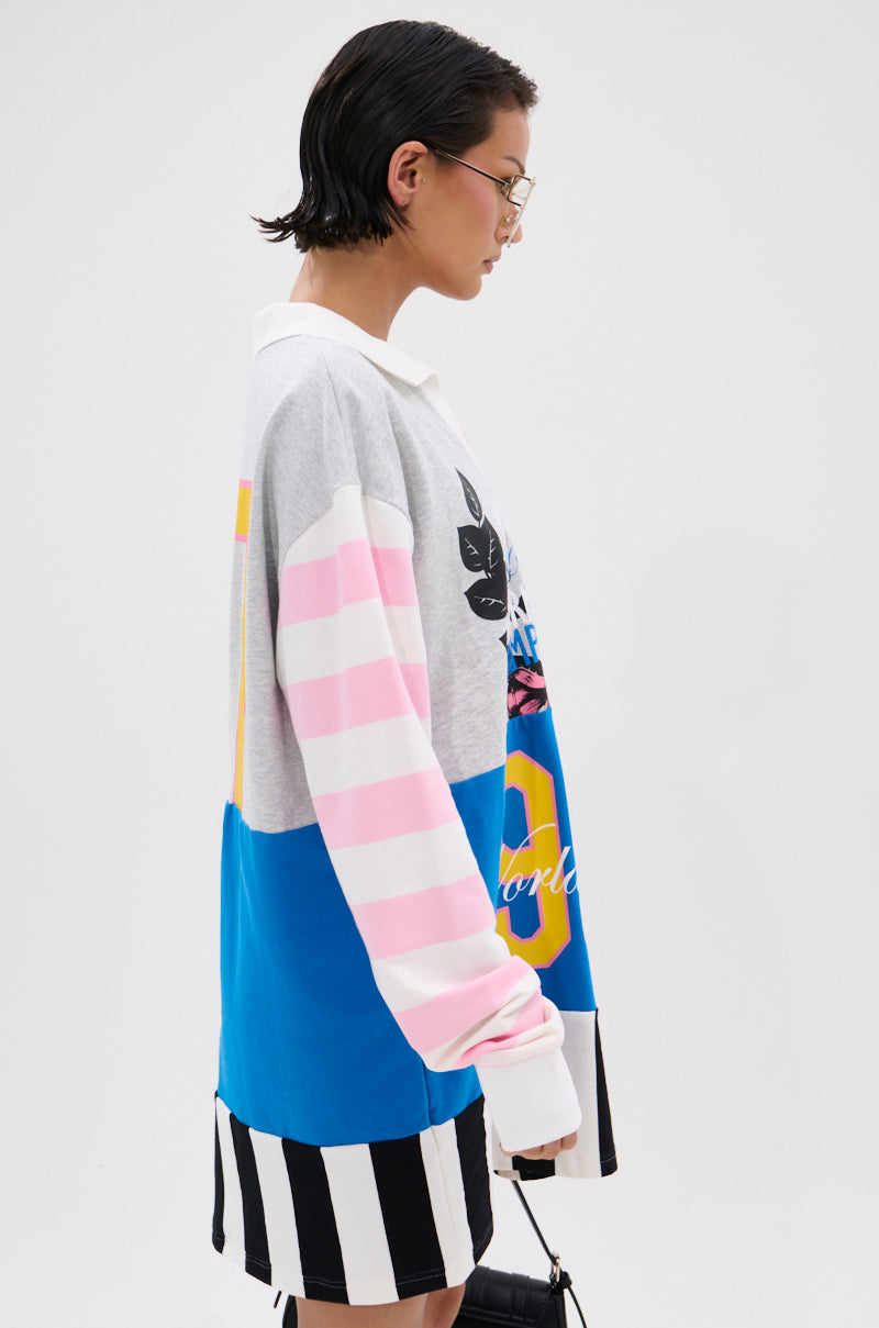 A person with short dark hair and glasses stands in profile, wearing the HERE FOR THE DRINKS PRINTED TSHIRT DRESS—a loose, colorful long-sleeve dress with colorblock stripes and varsity-inspired graphics.