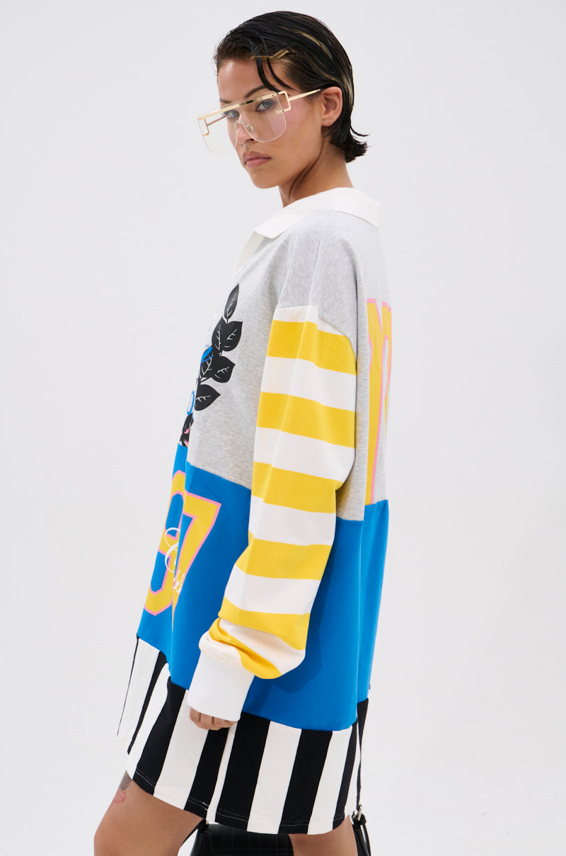 Person with short dark hair and glasses wears the HERE FOR THE DRINKS PRINTED TSHIRT DRESS—an oversized shirt with varsity graphics, yellow colorblock stripes, and bold blue accents—paired with black and white striped shorts against a white background.