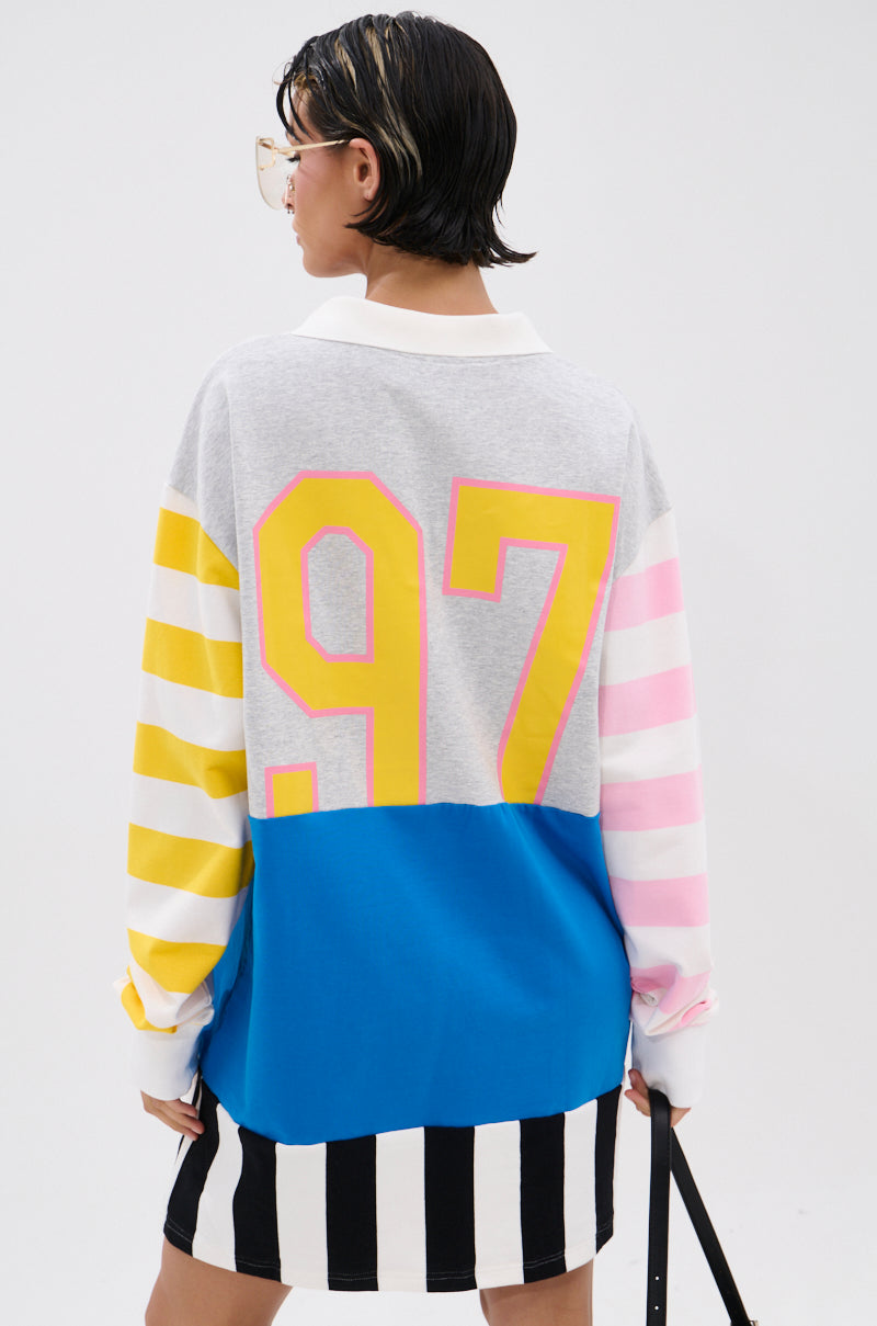 A person stands facing away, wearing the HERE FOR THE DRINKS PRINTED TSHIRT DRESS featuring yellow and pink striped sleeves, a large yellow "97" on the back, blue and gray panels, and a black-and-white striped skirt.