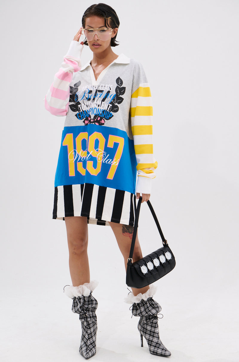 A person poses against a white background wearing the HERE FOR THE DRINKS PRINTED TSHIRT DRESS—an oversized, colorful T-shirt dress with varsity graphics and mixed patterns—paired with plaid boots, ruffle socks, visor sunglasses, and a black handbag.
