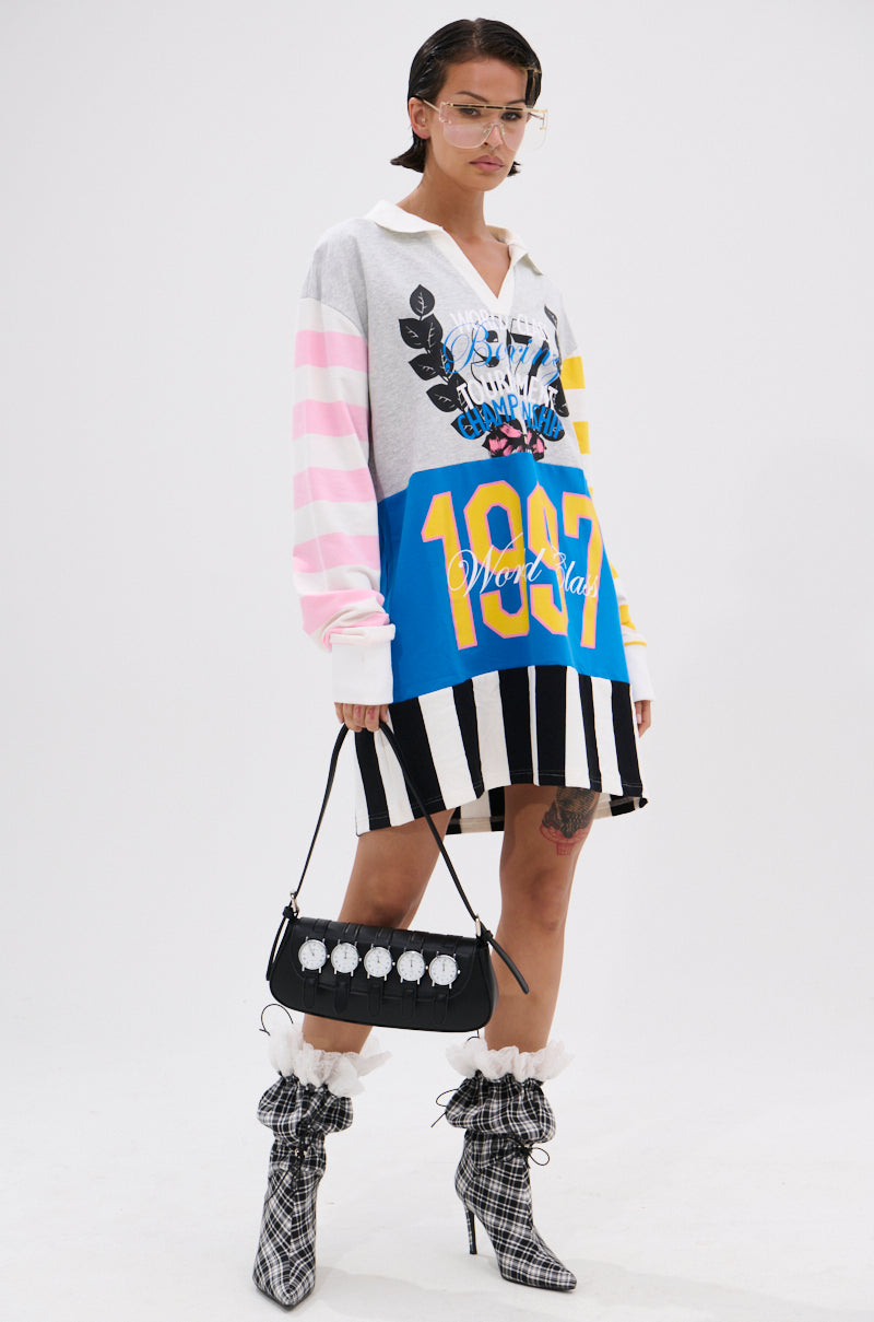 A person with short hair wears the HERE FOR THE DRINKS PRINTED TSHIRT DRESS and holds a black handbag, standing confidently against a plain white background.