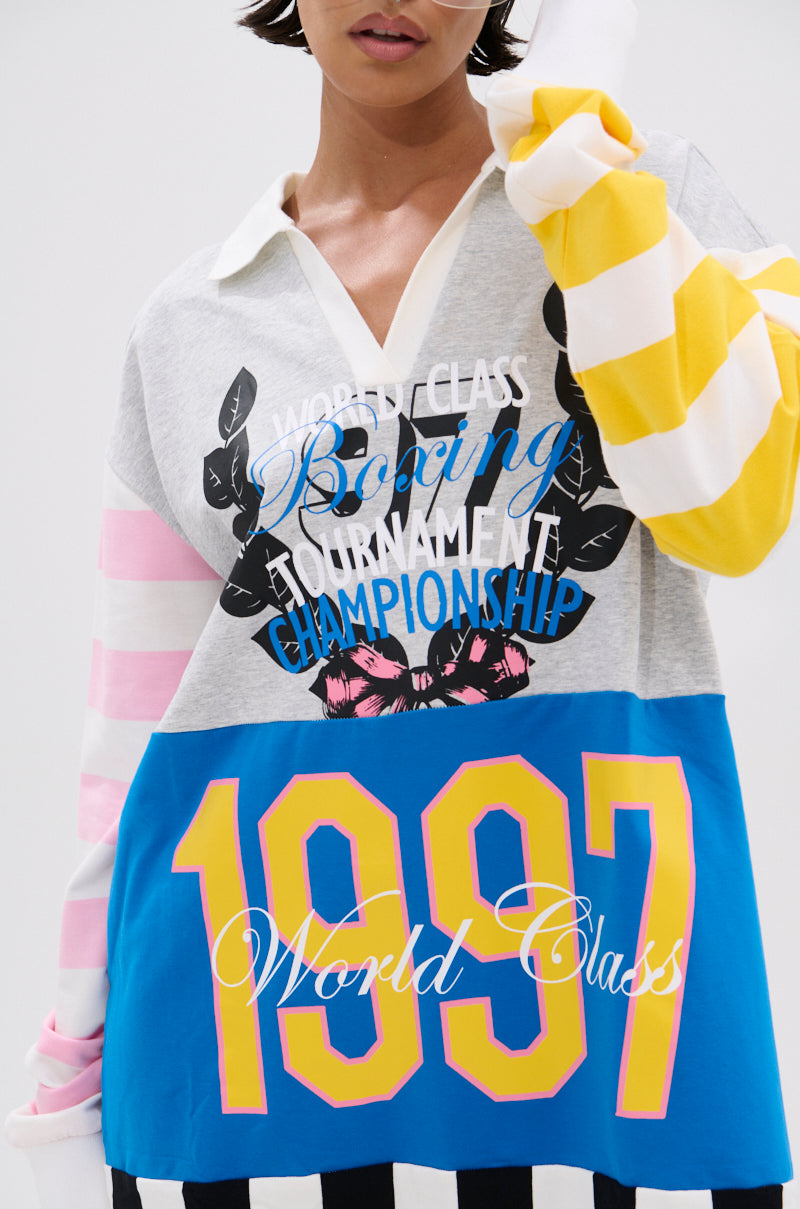 A woman wears the HERE FOR THE DRINKS PRINTED TSHIRT DRESS, an oversized style with colorblock yellow, white, pink stripes, blue and black sections, varsity graphics, the bold number "1997," and championship phrases.