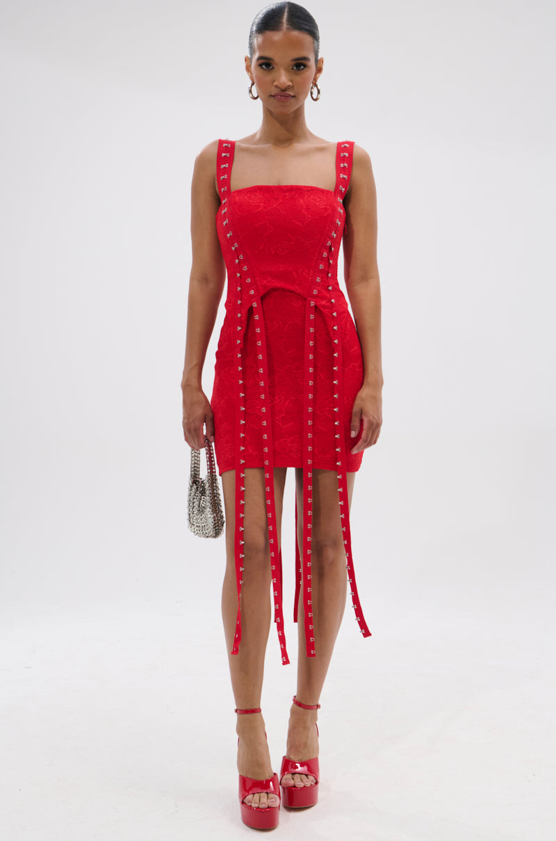 A woman stands against a white background wearing the I KNOW YOU MISS ME CORSETED LACE MINI DRESS IN RED, featuring a sleeveless design and square neckline. She pairs it with red platform heels and a small silver handbag.