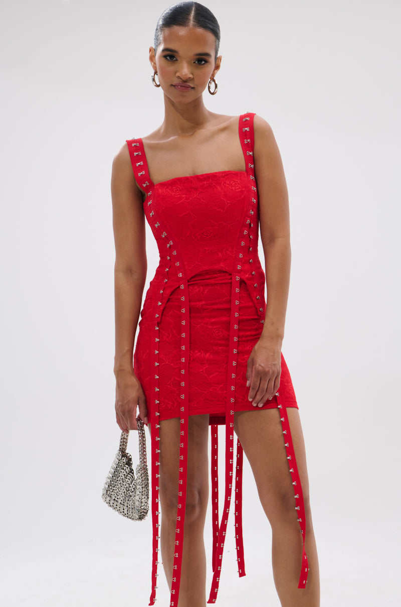 A woman poses against a white background wearing the I KNOW YOU MISS ME CORSETED LACE MINI DRESS IN RED, a fitted, sleeveless lace mini with decorative straps and metal studs, holding a small textured handbag.