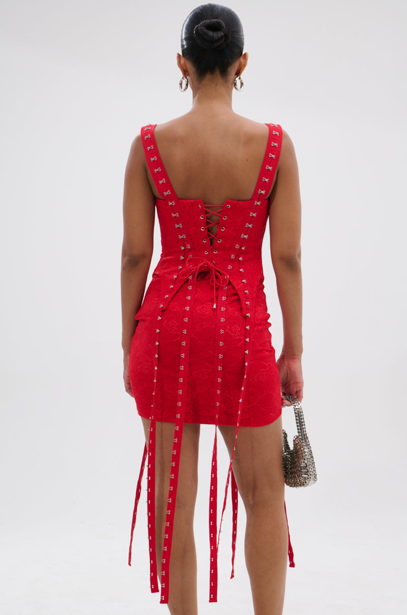 A woman faces away, wearing the I KNOW YOU MISS ME CORSETED LACE MINI DRESS IN RED with lace-up back and ribbon ties. She holds a silver handbag, her hair in a neat bun, set against a plain white background.