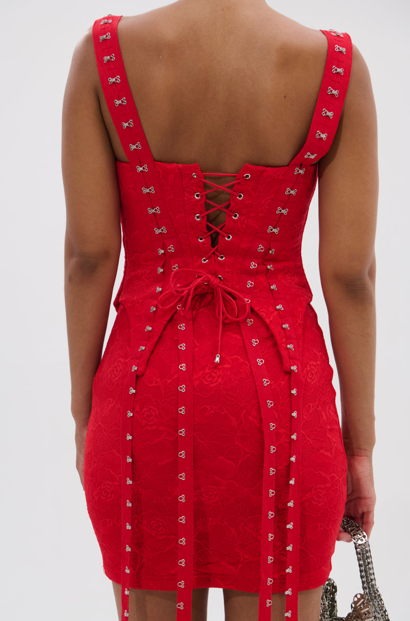 A woman faces away from the camera wearing the I KNOW YOU MISS ME CORSETED LACE MINI DRESS IN RED, featuring a fitted lace-up back, silver stud accents, and floral lace details. She holds a silver chain bag.