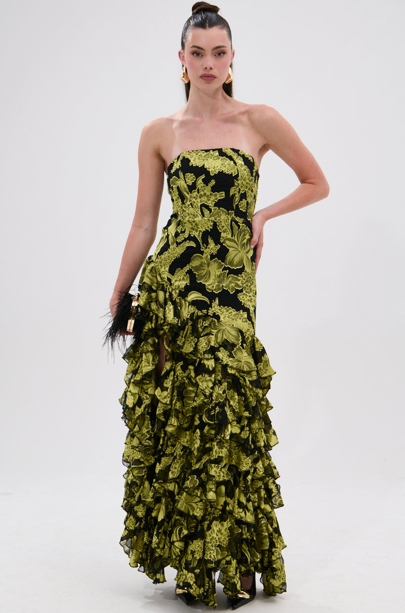 A woman poses in the STOLE THE SHOW SATIN RUFFLE MAXI DRESS, a strapless, floor-length gown with bold green floral print and cascading ruffled layers, accessorized with a black feather-detailed clutch.
