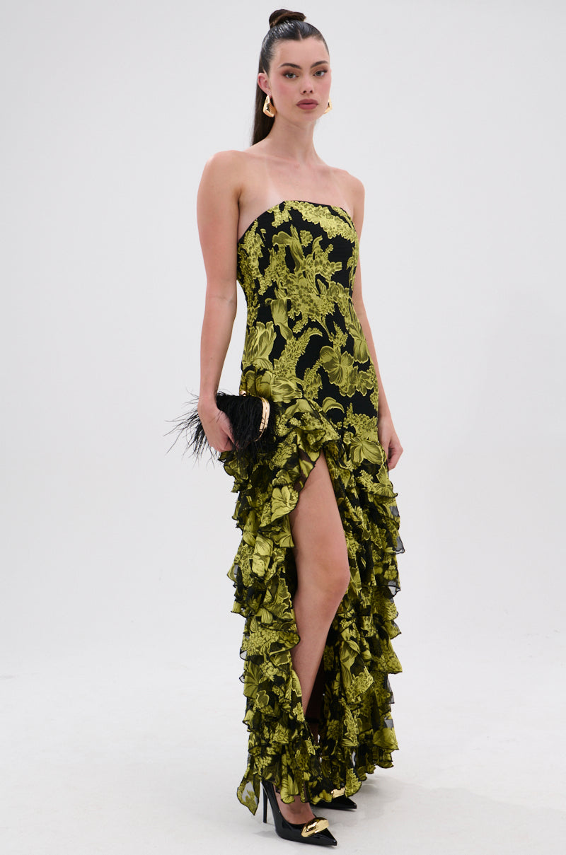 A woman stands against a plain white background wearing the STOLE THE SHOW SATIN RUFFLE MAXI DRESS in black and gold floral print with a thigh-high slit, paired with black heels, gold earrings, and a feathered black clutch.