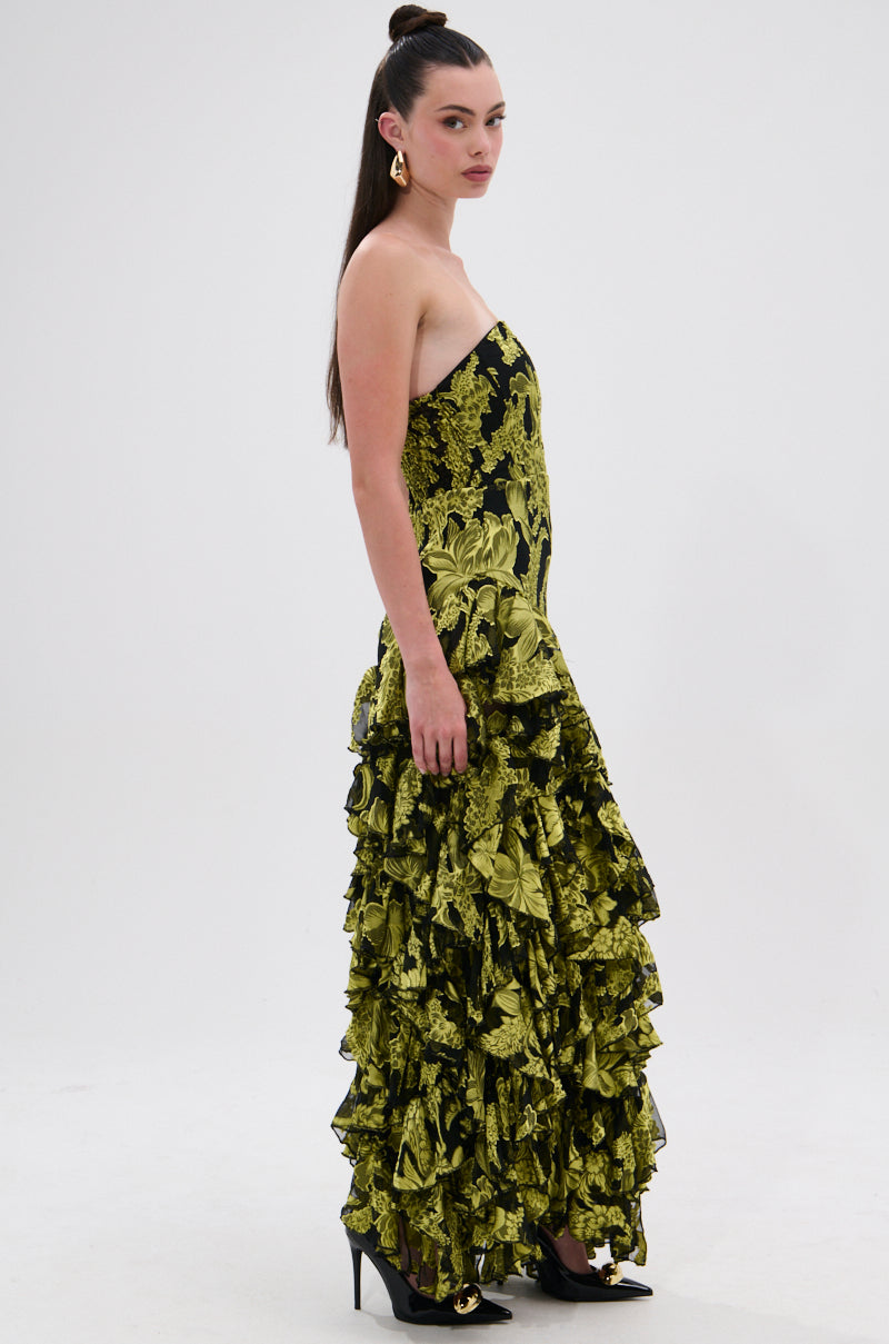 A woman with long dark hair stands sideways against a white background, wearing the STOLE THE SHOW SATIN RUFFLE MAXI DRESS in black and yellow floral print with layered ruffles, paired with black pointed-toe heels.