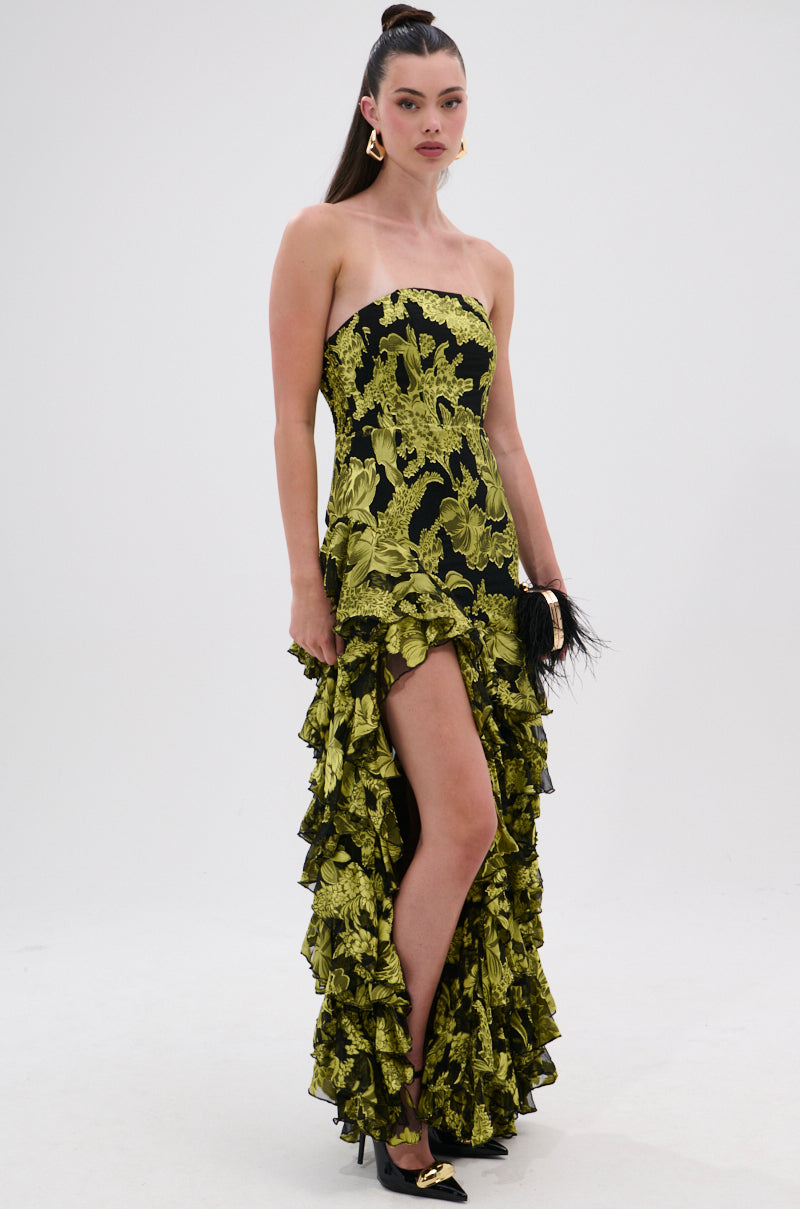 A woman poses in the STOLE THE SHOW SATIN RUFFLE MAXI DRESS, a strapless floral print maxi with bold green blooms on black satin, high slit, and ruffle details. She completes the look with black heels, gold earrings, and a sleek ponytail.