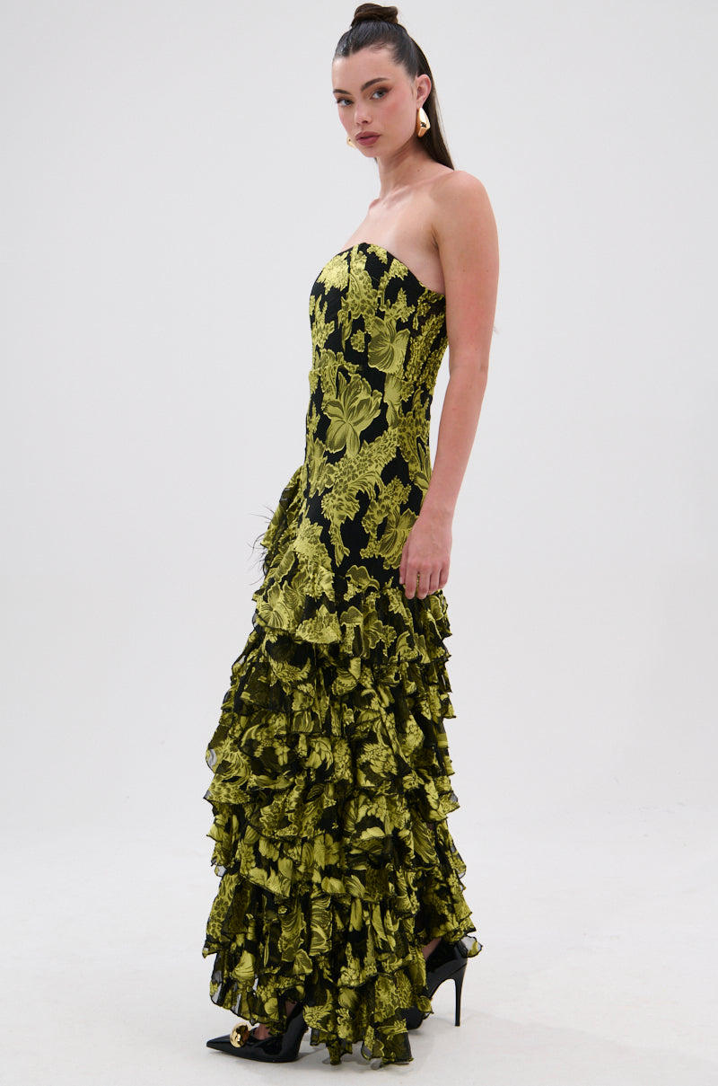 A woman models the STOLE THE SHOW SATIN RUFFLE MAXI DRESS—a strapless, floor-length black maxi dress with bold yellow floral patterns. She wears black heels, has long dark hair tied back, and poses against a plain white background.