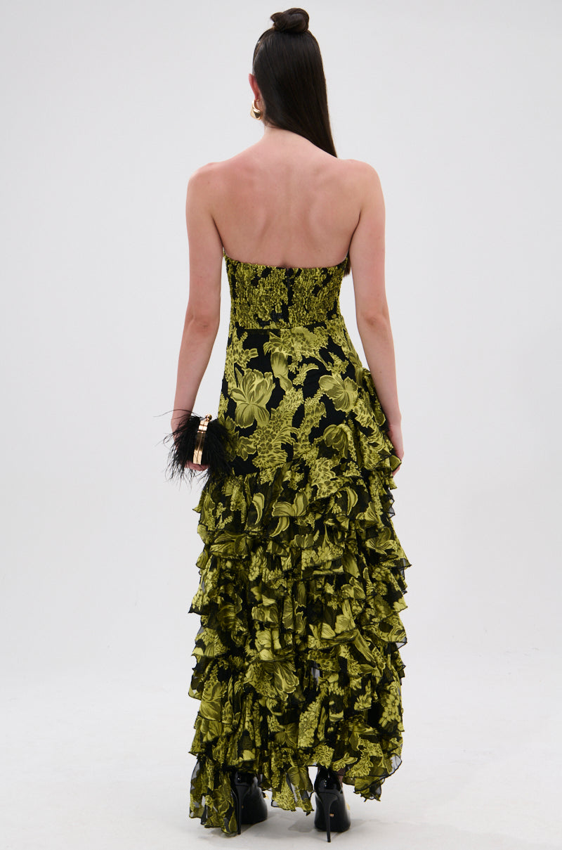A woman faces away, wearing the STOLE THE SHOW SATIN RUFFLE MAXI DRESS with layered ruffles. She holds a small black, feather-detailed clutch, and her long straight hair is styled half-up.
