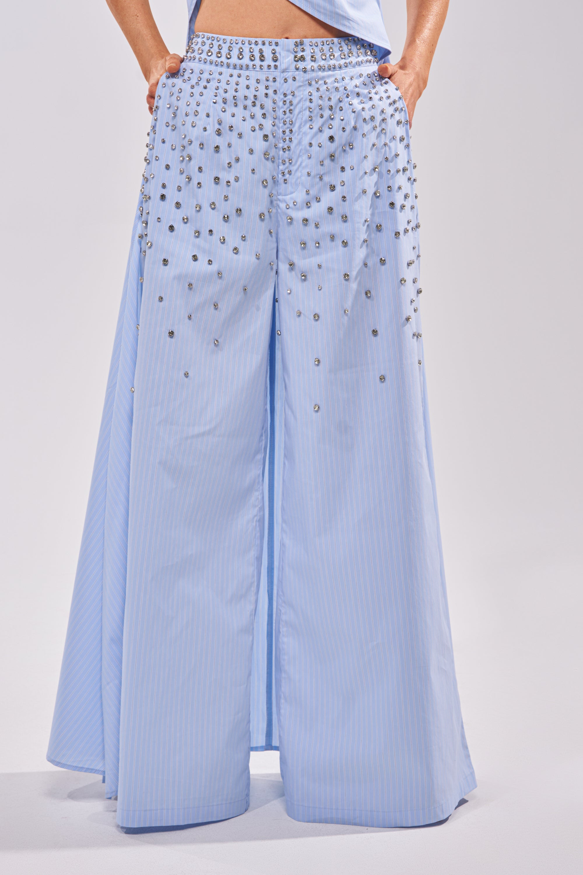 Wearing the STAR OF THE NIGHT POPLIN TROUSER in light blue, featuring a wide-leg silhouette and silver embellishments scattered from waist to upper thighs, a person stands with hands in their pockets. 0