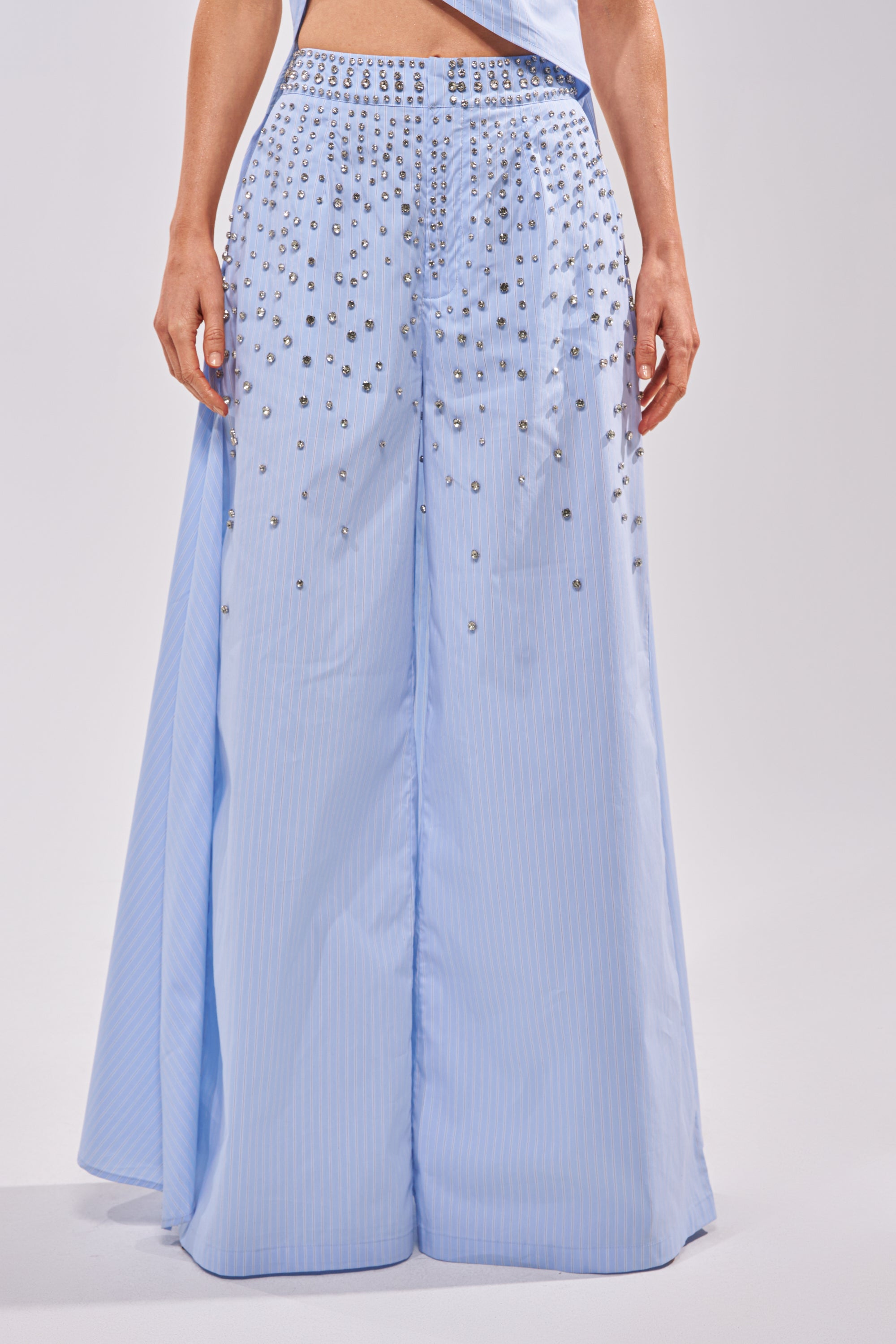 Someone stands against a plain light background wearing the STAR OF THE NIGHT POPLIN TROUSER, featuring a light blue color, wide-leg silhouette, vertical stripes, and delicate silver studs and beads. 1