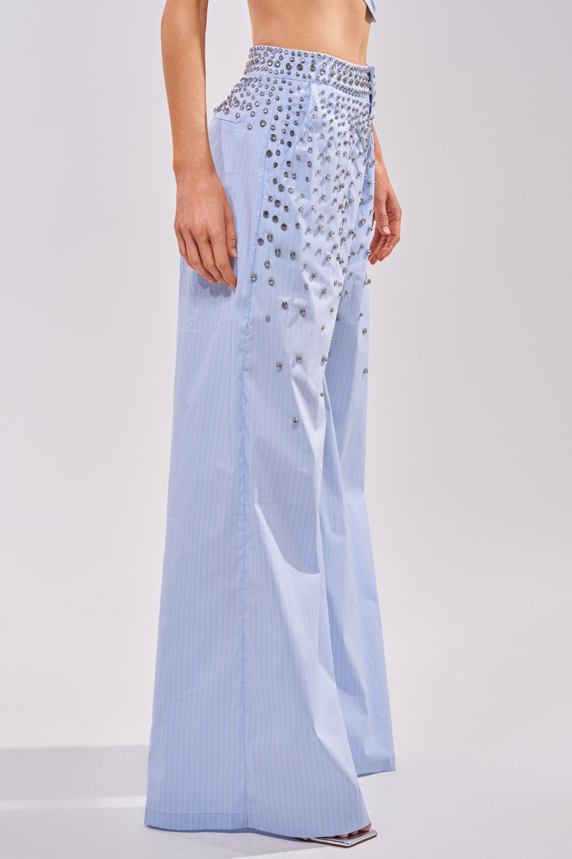Wearing the STAR OF THE NIGHT POPLIN TROUSER in light blue, this look features high-waisted, wide-leg trousers with subtle pinstripes and rhinestone embellishments clustered around the waist and upper thighs. 2