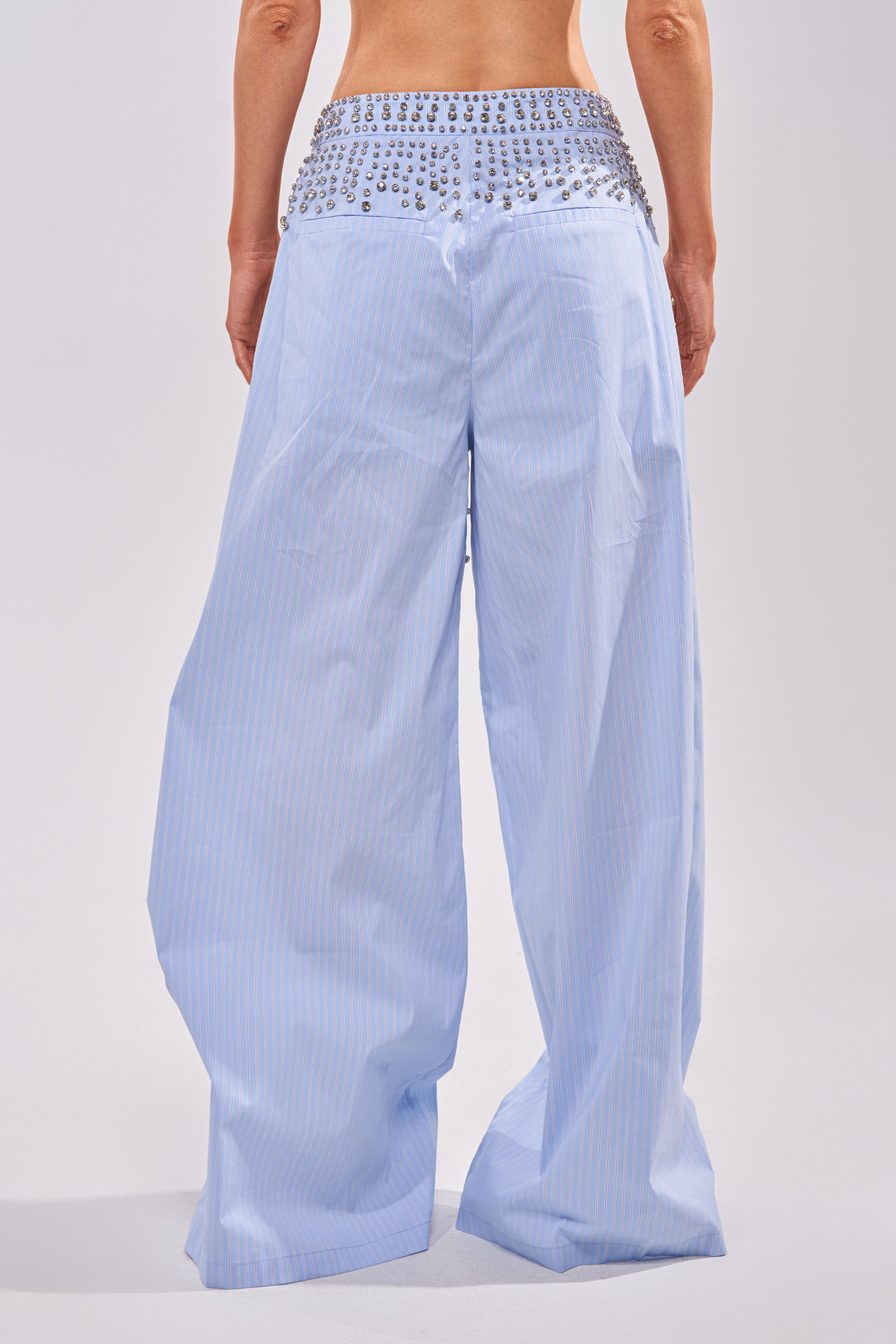 A person stands facing away, wearing the STAR OF THE NIGHT POPLIN TROUSER—light blue, high-waisted, wide-leg pants featuring subtle vertical stripes and sparkling rhinestone embellishments at the waistband. 3